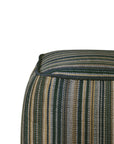 Close-up of a square Zahara Outdoor Pouf by Ethnicraft featuring a woven vertical stripe pattern in shades of green, yellow, brown, and cream with dark piping along the edge in weather-resistant fabric.