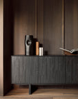 A modern interior with dark wood walls and floors features an Ethnicraft Mosaic Sideboard - 4 door holding a sculptural black vase, two cylindrical items, and an open book. The space showcases minimalist, elegant design.