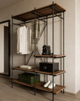 The Oscar Hanging Rack by Ethnicraft, crafted from FSC-certified teak and metal, displays hanging white shirts and a scarf. Its shelves hold a green suitcase, white sneakers, a black safe, and a coffee machine against a light wall and floor.
