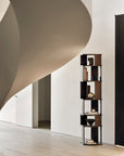 A modern, minimalist interior features the sculptural Stairs Column - Teak by Ethnicraft, a tall open bookshelf displaying books and decor, set against a plain white wall with light wooden flooring and a curved staircase.