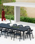A modern outdoor dining area showcases an Ethnicraft Bok Outdoor Dining Table in black teak with eight matching chairs on a white patio. A white vase with greenery sits atop the table as a person in red walks past lush plants and a beige wall.