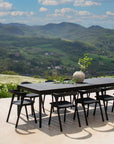 An Ethnicraft Bok Outdoor Dining Table in Black Teak, paired with six black chairs, sits on a patio overlooking hills and mountains. The table is topped with a vase of fresh green branches under a partly cloudy sky.