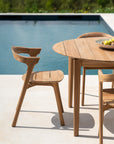 An Ethnicraft Bok Outdoor Round Dining Table in natural teak, paired with matching chairs, sits on a patio by the pool. A bowl of lemons decorates the table, with lush greenery in the background.