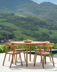 An Ethnicraft Bok Outdoor Round Dining Table in natural teak with four matching chairs sits on a patio overlooking green hills. A bowl of lemons decorates the center of the table.