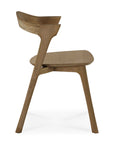 Side view of the Ethnicraft Bok Dining Chair - Natural Teak in FSC-certified solid teak, highlighting its modern sculptural design with a curved backrest and seat, angled legs, and minimalist style against a plain white background.