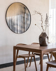 A modern dining area showcases an Ethnicraft wooden table with four Bok Dining Chairs in Natural Teak. On the FSC-certified solid teak tabletop rests a large brown vase with dried branches, while a round wall mirror reflects sheer curtains and soft light.