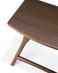 A close-up of the Ethnicraft Osso Dining Stool, crafted in FSC-certified teak with a smooth dark brown finish, showing part of the seat, one leg, and a support bar against a white background.