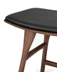 Close-up of the Ethnicraft Osso Dining Stool – Teak with Black Leather, showing its dark teak frame and black leather seat. Only part of the seat and two legs appear against a white background.