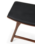 A close-up of the Ethnicraft Osso Dining Stool - Teak with Black Leather highlights its angled legs and smooth, minimalist teak design against a white background.
