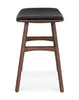 The Osso Dining Stool - Teak with Black Leather by Ethnicraft features a modern teak frame, curved supports, and a black leather cushioned seat, showcasing minimalist design and fine craftsmanship in front-view.