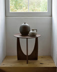 The Ethnicraft Geometric Side Table in varnished teak stands beneath a window, topped with two ceramic vessels, its wood glowing in natural light with greenery outside.