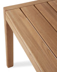 Close-up of the Ethnicraft Jack Outdoor Dining Table - Teak, featuring smooth, light brown solid teak with evenly spaced slats on the tabletop and sturdy, angled legs.