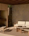 A modern outdoor patio showcases an Ethnicraft Jack Woven Outdoor Lounge Chair and light wood sofa with neutral cushions, complemented by matching coffee tables beneath a wooden roof and set against a textured stone wall.