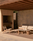 A modern patio with a wooden roof showcases a cushioned sofa, woven lounge chair, and two Ethnicraft Boomerang Outdoor Coffee Tables with hand-finished concrete tops on a brown rug. A built-in grill stands against a stone-textured wall.
