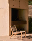 An Ethnicraft Jack Woven Outdoor Lounge Chair with an Italian weave seat and backrest sits on a patio before a modern outdoor fireplace in a beige concrete wall, accented by greenery and a wooden screen for added warmth.
