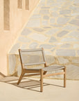 The Jack Woven Outdoor Lounge Chair by Ethnicraft, with an Italian weave seat and backrest, sits on a sunlit patio beside a stone wall and beige stucco, casting a soft shadow on the ground.