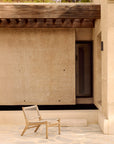 An Ethnicraft Jack Woven Outdoor Lounge Chair sits on a stone patio before a beige stucco building with a wooden pergola and glass door, creating a minimalist, serene outdoor setting.