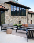 A modern patio with gray cushioned seating, coffee table, and beige pouf atop the Ethnicraft Nomad Outdoor Rug, made from recycled PET yarns, set against a stone house with large windows and terracotta roof.
