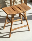 The Ethnicraft Osso Outdoor Stool, featuring a smooth teak finish, two rectangular seat slots, and angled legs, is shown on a light floor in sunlight.