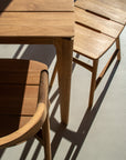 Two wooden chairs, an Ethnicraft Osso Outdoor Stool, and a wooden table with smooth, light-brown finishes sit on a sunlit floor. Shadows from the Ethnicraft outdoor collection cast diagonal lines across the scene.