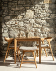 An Ethnicraft outdoor wooden table with three cushioned chairs, a bench, and the Osso Outdoor Stool sits in sunlight by a rustic stone wall; a single beverage glass with a straw rests on the table.
