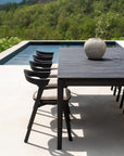 The Ethnicraft Bok Outdoor Dining Table in black teak with four black chairs sits by the pool, adorned with a ceramic vase and green plant. Lush greenery and trees create a serene backdrop.