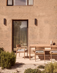A minimalist outdoor patio showcases a solid teak dining table with three chairs, including a nearby Jack Outdoor Dining Chair - Teak (without armrest) by Ethnicraft, all surrounded by greenery in front of a textured beige wall with a glass door and window.