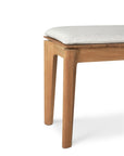 A close-up of the Ethnicraft Bok Outdoor Bench - Natural Teak, featuring a light-upholstered seat cushion, solid teak construction, two legs, and a corner detail shown against a white background.