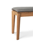 A close-up of the Ethnicraft Bok Outdoor Bench - Natural Teak, featuring solid teak construction, angled legs, and a textured gray cushioned seat set against a white background.