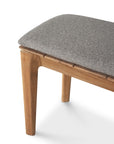 Close-up of the Ethnicraft Bok Outdoor Bench - Natural Teak, featuring angled legs and a cushioned gray fabric seat, crafted from solid teak and shown against a white background.