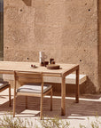 An Ethnicraft Jack Outdoor Dining Table in solid teak with two chairs sits by a textured beige wall, set with stacked plates, cups, a white pitcher, and a small dish as sunlight creates shadows on the ground.