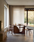 A minimalist dining area features the Ethnicraft Geometric Dining Table in brown teak, paired with four wooden chairs and simple glassware. Large windows with sheer curtains overlook a garden, while neutral tones create a calm atmosphere.