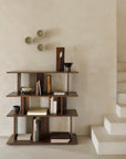 A minimalist room features the Ethnicraft Irregular Rack - Teak, crafted from FSC-certified wood, displaying books and decor against a beige wall. Above are three round wall sconces, with white stairs on the right.