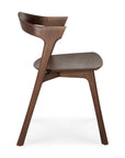 Side view of the Ethnicraft Bok Dining Chair - Brown Teak, crafted from FSC-certified teak with a curved backrest and angled legs. This modern chair features a smooth, minimalist design and rich brown finish against a white background.