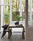 A modern dining area showcases the Ethnicraft Bok Dining Table in brown teak, complemented by black chairs and minimalist tableware. Tall windows framed by greenery fill the space with natural light.