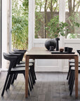 A modern dining room showcases the Ethnicraft Bok Dining Table in brown teak, paired with six black chairs. Large windows frame lush greenery and fill the space with natural light.