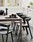 A modern dining area showcases an Ethnicraft Bok Dining Table in brown teak paired with black chairs, minimalist tableware, and white walls with large windows. A plant and bowl rest on a white countertop in the background.