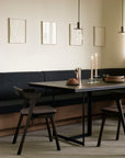 A modern dining space showcases an Ethnicraft Tacet Dining Table, dark wood chairs, a bench, two glowing candles, a loaf of bread, minimalist art on a light wall, and elegant pendant lights suspended above.