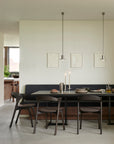 A modern dining area showcases the Ethnicraft Tacet Dining Table with a teak top, four dark chairs, and a built-in bench. Two pendant lights hang above the set table, while minimalist art decorates the light-colored wall.