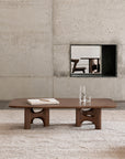 Low rectangular Ethnicraft Flow Coffee Table | Teak Wild Brown with solid wood construction and four curved, arch-shaped legs, holding a book, a tall glass, and a small glass.