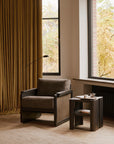 An Ethnicraft Panel Lounge Chair - Dark Brown Teak sits by a window with mustard curtains, next to a wooden side table holding a teapot, cup, and book. Autumn trees can be seen outside.