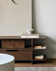 The Ethnicraft Kabuki TV Cupboard, crafted from FSC-certified teak, displays a vintage box, books, and a white ceramic bowl on its shelves. Minimalist art sits on top, while a round white table is partly visible in the foreground.