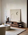 A minimalist living room featuring the Ethnicraft Kabuki TV Cupboard in dark wood with books and decor, a large abstract art piece, round white coffee table, beige rug, light armchair, and a tall branch in a slim vase.