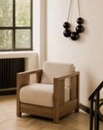 The Roan Lounge Chair | Flax by Ethnicraft, with a wooden frame and beige cushions, sits in a bright room featuring large windows, light wood floors, and wall art with black hanging spheres.
