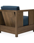 A modern, vintage-inspired Roan Lounge Chair | Ink by Ethnicraft, featuring a wooden frame and blue upholstered cushions, is shown from a rear-side angle. The back displays an angled wood panel for support, characteristic of the Roan Lounge Chair’s distinctive design.