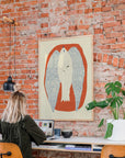 A blonde graphic designer works at a desk in front of a brick wall, with PSTR Studio’s Olivia Leino - Two open shells under the Sun (Framed Print) hanging above and a potted plant nearby.