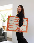 A woman with curly hair in dark clothing smiles while holding the Blooming Season framed print by Olivia Leino for PSTR Studio, featuring white shapes on orange, showcased in a modern living room with plants and books.