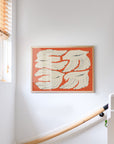 The Olivia Leino - Blooming Season framed print by PSTR Studio, featuring cream bird-like shapes on an orange background, hangs beside a staircase with a wooden handrail and small window with wooden blinds.
