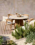 The Roller Max Outdoor Dining Table by Ethnicraft, with a light top and reclaimed teak base, is paired with three wooden chairs with gray cushions on a patio surrounded by greenery and a stone wall backdrop.