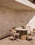 A minimalist outdoor dining area features Ethnicrafts Roller Max Outdoor Dining Table, three wooden chairs, and a decorative stone bowl, set against a textured stone wall and partially shaded by a concrete overhang.